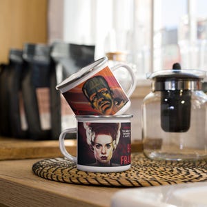 May include: Two white enamel mugs stacked on a woven mat. The top mug features a portrait of Frankenstein's monster, while the bottom mug displays the Bride of Frankenstein. The mugs have silver rims and white handles. The background includes coffee bags and a glass teapot.