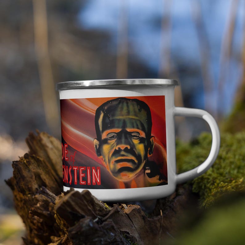 May include: White enamel mug featuring a vintage illustration of Frankenstein's monster. The artwork is set against a red and orange background, with the name "Frankenstein" in red text. The mug has a silver rim and a white handle.