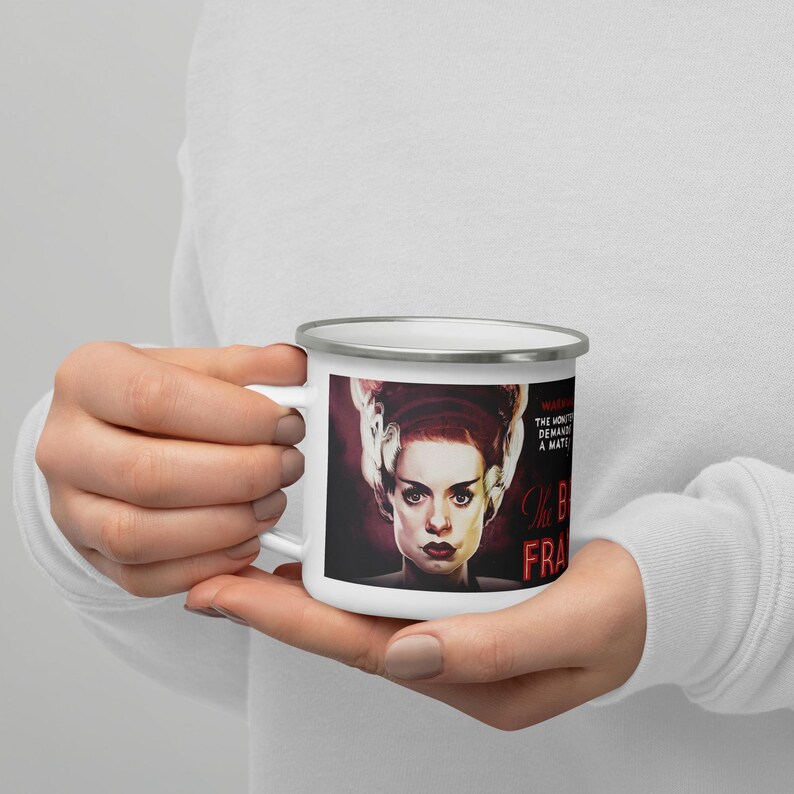 May include: White enamel mug featuring a portrait of the Bride of Frankenstein with the text "The Bride of Frankenstein". The mug has a silver rim and a white handle. The background is a dark red.