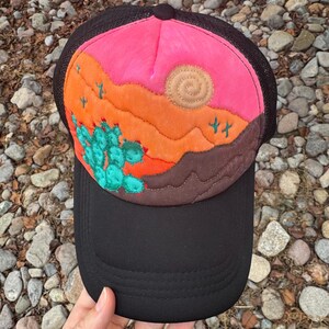 May include: Black trucker hat with a colorful desert scene. The hat features a pink and orange sunset, brown mountains, a turquoise cactus, and a tan sun. The brim is black, and the mesh backing is dark brown.