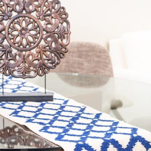 May include: A decorative, round, bronze-colored wall hanging with intricate floral and scrollwork stands on a black stand. A white and blue geometric patterned table runner with fringe is on a glass table.