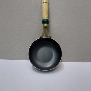 Puede incluir: Pequeña sartén redonda negra con mango de madera. El mango es de color marrón claro con un acento verde cerca de la sartén. La sartén tiene un lazo de metal en el extremo del mango para colgar. Utensilios de cocina.