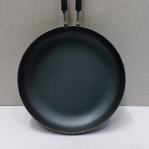 Puede incluir: Una sartén negra y redonda con un mango largo negro y un lazo de metal plateado para colgar. La sartén está hecha de un material oscuro y no reflectante, para cocinar.