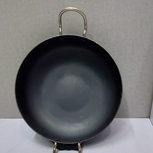 May include: A black wok-style pan with two metal handles. The pan has a rounded shape and a dark interior. The handles are attached to the sides of the pan and are curved. The pan is designed for cooking.