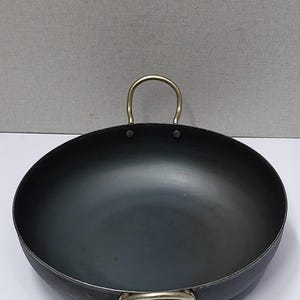 May include: A black wok-style pan with a rounded base and two brass-colored handles. The pan has a wide, open design, suitable for stir-frying and other cooking methods. The handles are riveted to the pan's body.