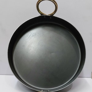 Peut inclure: Une po&ecirc;le ronde en m&eacute;tal gris fonc&eacute; avec deux poign&eacute;es en anneau de couleur or. La po&ecirc;le a un design large et peu profond, adapt&eacute; &agrave; la cuisson ou au service. Les poign&eacute;es sont fix&eacute;es au bord de la po&ecirc;le.