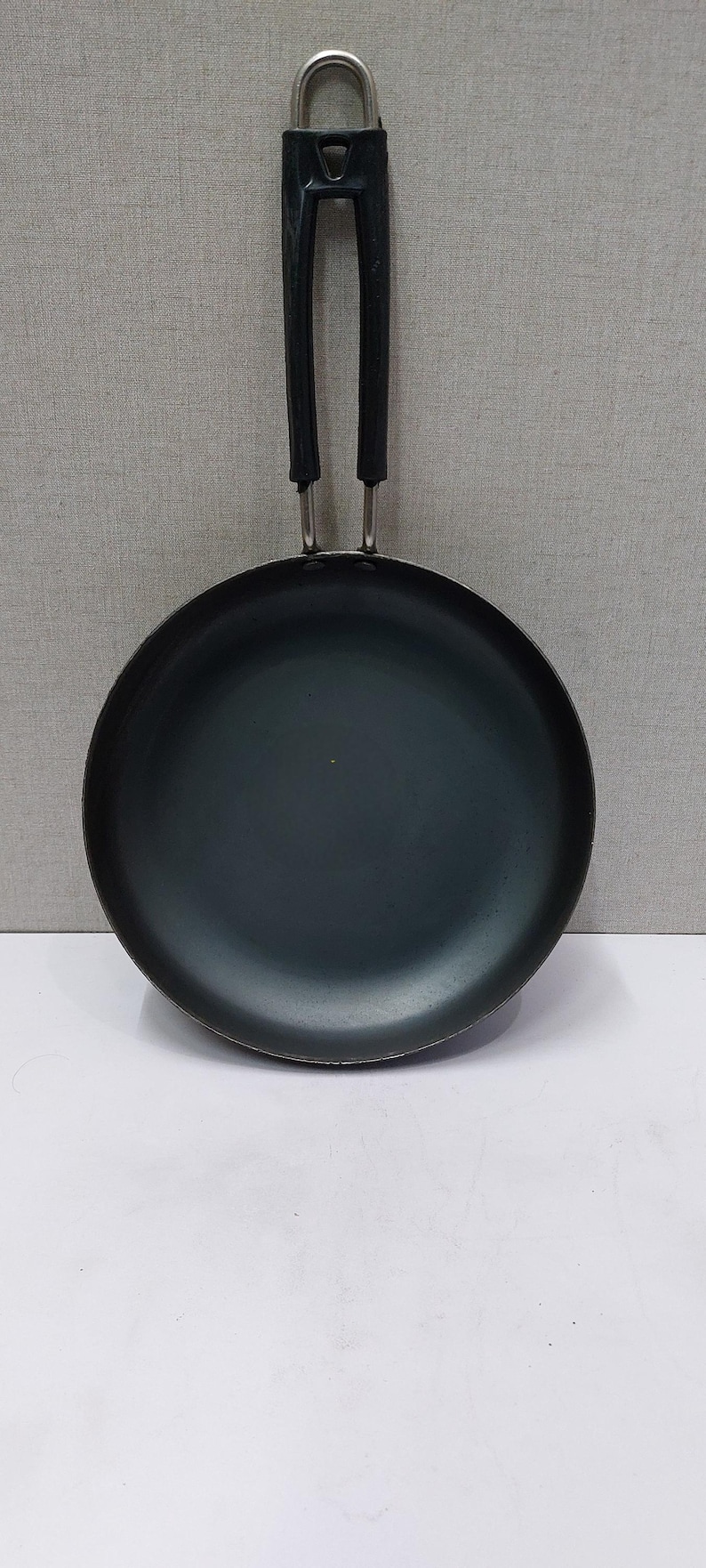 May include: A black, round frying pan with a long, black handle and a silver metal loop for hanging. The pan is made of a dark, non-reflective material and is designed for cooking.