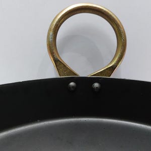 May include: Close-up of a black cooking pan with a gold-colored handle. The handle is a large, circular loop attached to the pan with two rivets. The pan's interior is dark, and the handle shows signs of wear.