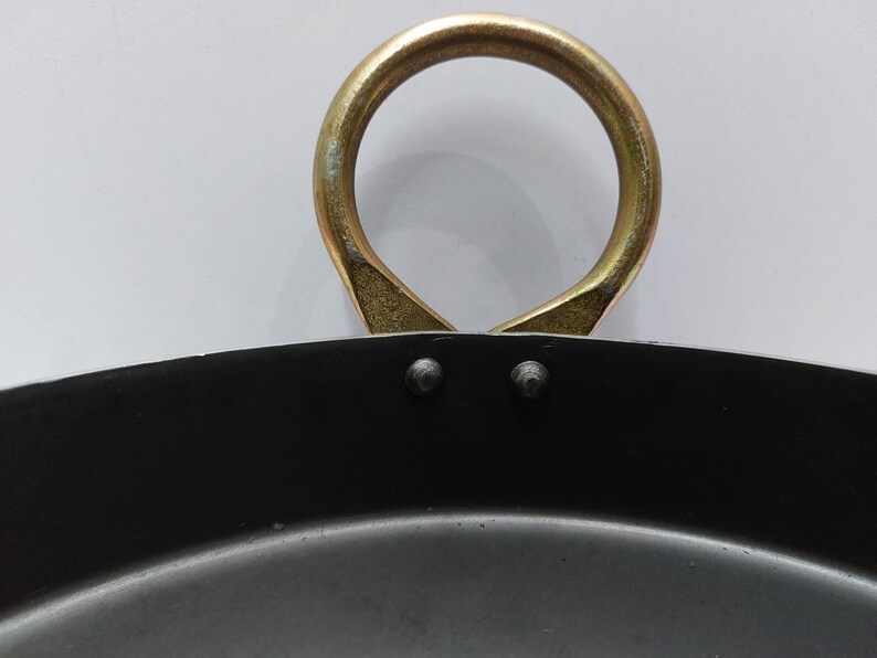 May include: Close-up of a black cooking pan with a gold-colored handle. The handle is a large, circular loop attached to the pan with two rivets. The pan's interior is dark, and the handle shows signs of wear.