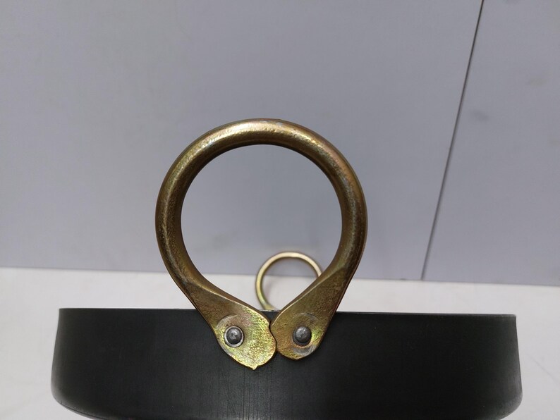 May include: Close-up of a black metal pot with a large, gold-colored handle. The handle is a rounded ring attached to the pot with two gold-colored brackets. A smaller gold ring is visible in the background.
