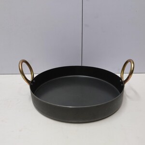 May include: A round, black metal pan with two brass handles. The pan has a shallow depth and a wide, flat base. The handles are arched and attached to the sides of the pan. The pan is likely used for cooking or serving food.