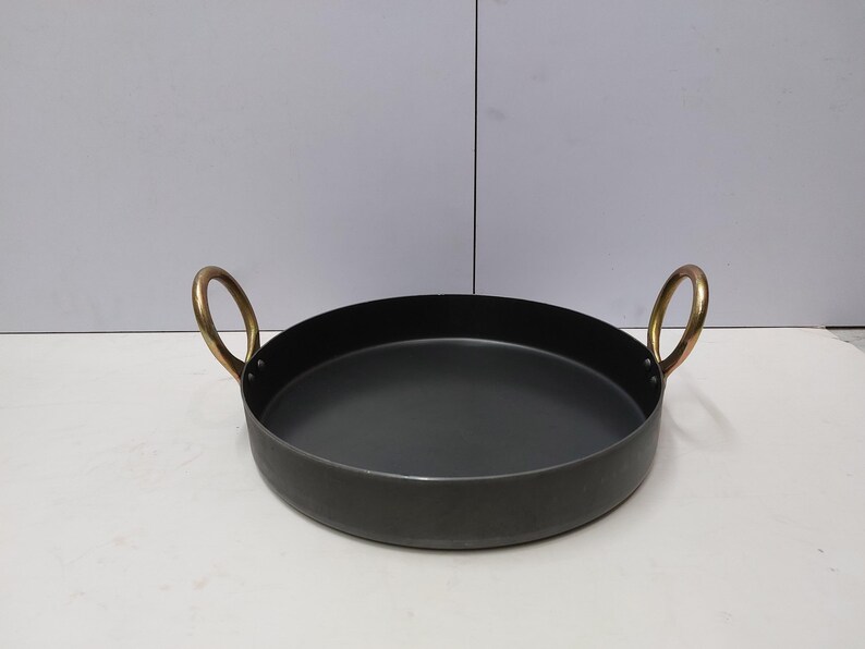 May include: A round, black metal pan with two brass handles. The pan has a shallow depth and a wide, flat base. The handles are arched and attached to the sides of the pan. The pan is likely used for cooking or serving food.