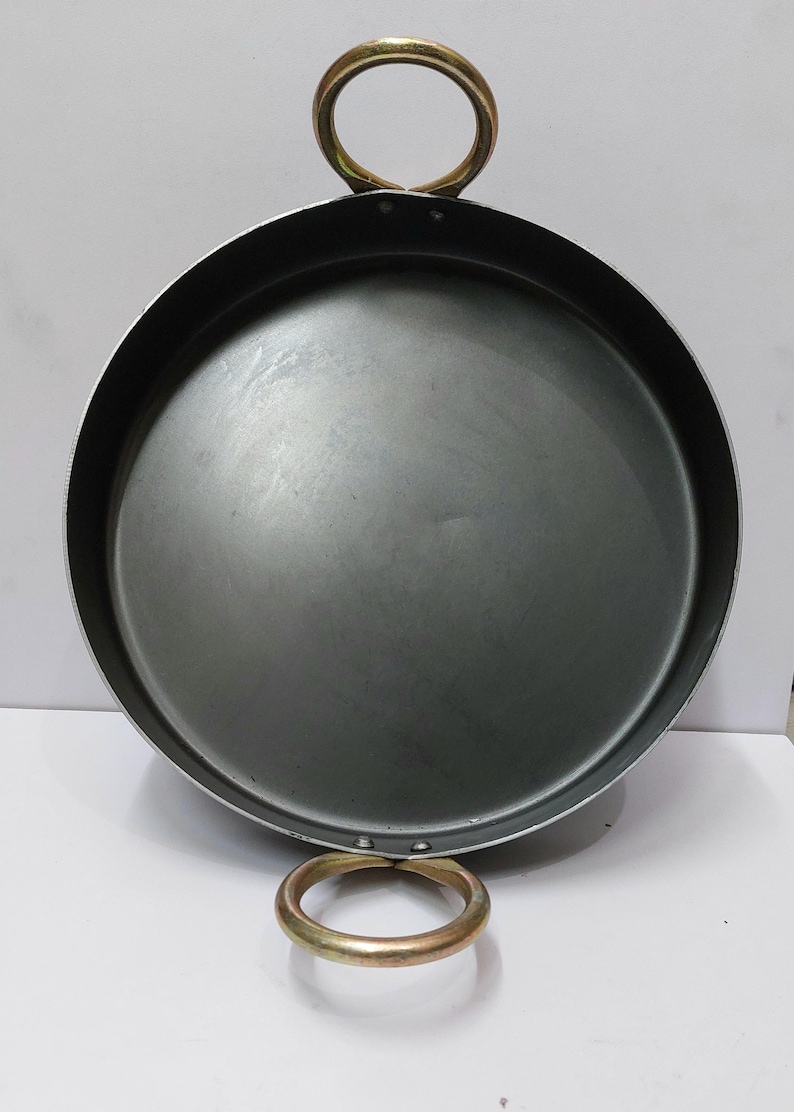 Peut inclure: Une po&ecirc;le ronde en m&eacute;tal gris fonc&eacute; avec deux poign&eacute;es en anneau de couleur or. La po&ecirc;le a un design large et peu profond, adapt&eacute; &agrave; la cuisson ou au service. Les poign&eacute;es sont fix&eacute;es au bord de la po&ecirc;le.