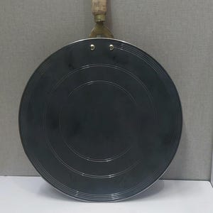 May include: A round, flat, dark gray metal griddle pan featuring a series of concentric circular grooves on its surface. The pan is attached to a short, cylindrical wooden handle by a brass-colored metal bracket secured with two silver rivets.