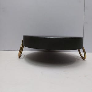 May include: A black, round footstool with a padded top and three gold-colored, ring-shaped legs. The footstool is a low, circular design, suitable for seating or as a decorative accent piece. The overall aesthetic is simple and elegant.