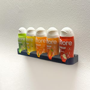 Könnte beinhalten: Fünf bunte Sirupflaschen in einem schwarzen Halter vor einer weißen Wand. Die Flaschen sind grün, gelb, orange und rot, jede mit einem weißen Deckel. Auf den Flaschen steht das Wort "more".