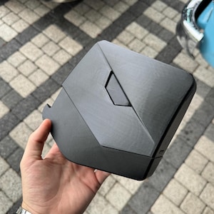 Könnte beinhalten: Ein mattschwarzes, geometrisch geformtes Objekt mit einem vertieften rautenförmigen Detail. Das Objekt hat eine strukturierte Oberfläche und scheint aus einem starren Material zu bestehen. Der Artikel wird in der Hand einer Person gehalten, um seine Größe und Form zu zeigen.
