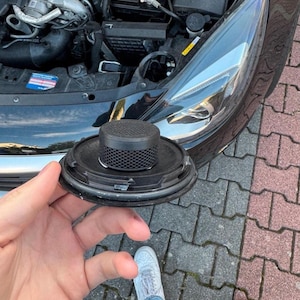 Könnte beinhalten: Eine schwarze Autolautsprecherkomponente mit einem Mesh-Top und einer kreisförmigen Basis wird vor der geöffneten Motorhaube eines Autos gehalten. Der Lautsprecher ist schwarz und hat eine strukturierte Oberfläche. Der Autoscheinwerfer ist sichtbar.