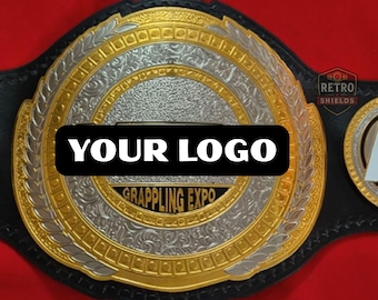 May include: A championship belt featuring a large circular gold and silver metal plate with intricate scrollwork and laurel leaf borders. The center displays a black rectangular plaque with the white text YOUR LOGO and the text GRAPPLING EXPO.