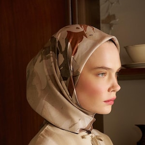 Puede incluir: Una mujer con un pañuelo beige con estampado floral, combinado con un conjunto beige a juego. El pañuelo cae elegantemente y el conjunto tiene un cierre de botones. El fondo incluye un armario de madera.