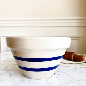May include: Cream-colored ceramic mixing bowl with two bold blue stripes. The bowl has a wide rim and a slightly tapered shape. A wooden rolling pin rests on a linen cloth in the background. Ideal for baking and kitchen decor.