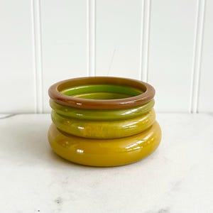 May include: A stack of five colorful bangles in shades of brown, green, and yellow. The bangles are made of a glossy material and are stacked on top of each other. The colors range from a deep brown to a bright yellow.