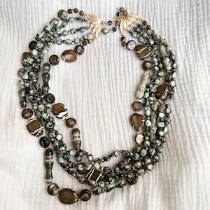 May include: A multi-strand necklace featuring a collection of polished beads in shades of dark brown, charcoal, and pale seafoam green. The strands are connected by two gold-toned metal leaf-shaped clasps at the top, creating a layered design.