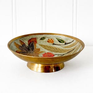 May include: A vintage brass bowl with a pedestal base. The interior features a floral design with brown, orange, and green accents on a light green background. The rim and base are a shiny gold color.