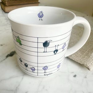 May include: White ceramic mug with a whimsical design of cartoon birds perched on musical staff lines. The birds are in various colors, including green, purple, and black. The mug has a curved handle and is suitable for hot beverages.
