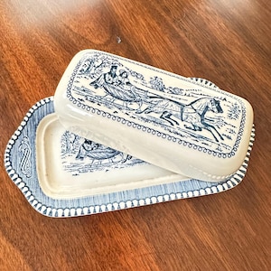 May include: A cream-colored ceramic butter dish with a rectangular lid and base. The dish features a blue transferware pattern depicting a horse-drawn sleigh with two figures. The border of the lid and base is decorated with a blue scrollwork design.