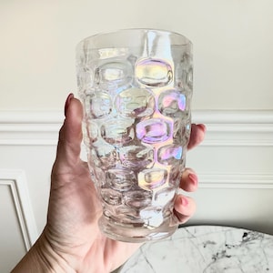May include: A clear, iridescent glass with a textured, bubble-like pattern. The glass is held in a hand, showcasing its unique design and the play of light on its surface. The glass is a drinking glass.