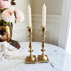 May include: Two vintage brass candlesticks with white candles. The candlesticks have a square base and a detailed, ornate design. The candles are tall and slender, and the overall aesthetic is elegant and classic.