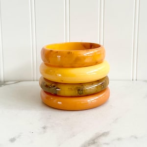 May include: A stack of four vintage bangles in shades of orange, yellow, and brown. The bangles are made of a glossy material and have a smooth, rounded shape. The colors range from a deep amber to a pale yellow.