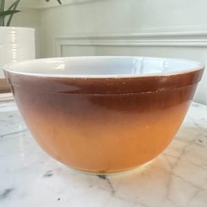 May include: A round mixing bowl with a smooth, glossy finish, featuring a color gradient that transitions from a dark brown rim at the top to a lighter, warm orange hue at the base, with a clean white interior surface visible from the top.