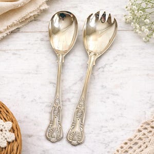 May include: A pair of silver serving utensils, a spoon and a fork, with ornate floral detailing on the handles. The spoon has a rounded bowl, while the fork has three tines. The utensils are displayed on a light-colored surface.