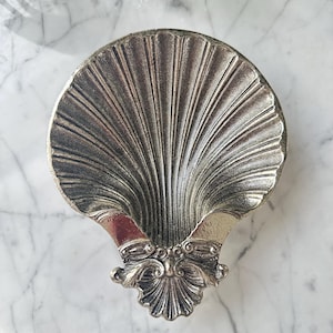 May include: A silver-toned metal dish shaped like a scallop shell. The interior features deep, radiating grooves extending from the base to the outer edge. The bottom hinge area displays intricate, raised scrollwork and decorative relief detailing.