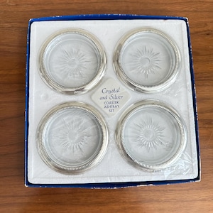 May include: A set of four clear glass coasters with a starburst design, each encased in a silver-colored metal rim. The coasters are presented in a blue and white box, with text that reads "Crystal and Silver COASTER ASHTRAY SET."