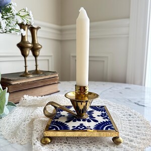 May include: A brass candlestick with a white candle. The base is a square tile with a blue and white floral pattern. The candlestick has a handle and sits on four small brass feet. Decorative home decor.