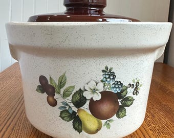May include: A cream-colored ceramic casserole dish with a speckled finish and a dark brown lid. The front features a botanical illustration of a yellow pear, a brown round fruit, green leaves, a white flower, and clusters of blue and dark berries.
