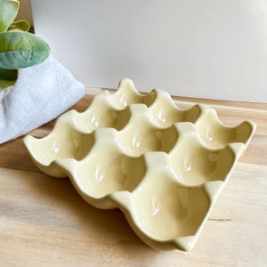 May include: A cream-colored ceramic egg holder with a wavy design, holding space for a dozen eggs. The egg tray is on a light wooden surface, with a white cloth and green plant in the background.