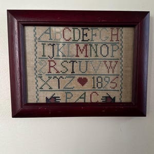 May include: Framed cross-stitch sampler with a burgundy wooden frame. The sampler features the alphabet in various colors, a heart, the number 1895, and the word PAC. The background is a tan fabric.