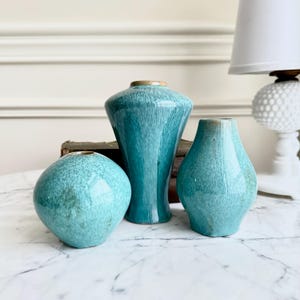 May include: Three turquoise ceramic vases of varying shapes and sizes. The vases have a speckled texture and a glossy finish. The tallest vase is a flared shape, while the other two are rounded. The vases are displayed on a marble surface.