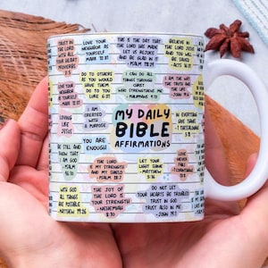 May include: White ceramic mug with a wraparound design featuring the text "My Daily Bible Affirmations" and various Bible verses. The mug is held by a person. The mug is a religious item.