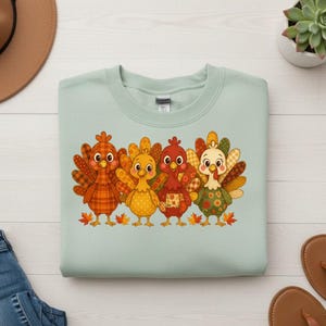 Könnte beinhalten: Hellblaues Sweatshirt mit einer Grafik von vier Zeichentrick-Truthähnen mit bunten Federn und Herbstblättern. Die Truthähne sind in Orange-, Gelb- und Rottönen gehalten. Ein saisonaler Artikel für Herbst oder Thanksgiving.