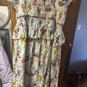 May include: A white dress with a floral pattern featuring birds, flowers, and fruit. The dress has a smocked bodice and a tiered skirt. The colors include yellow, orange, green, and blue. The dress has short sleeves and a midi length.