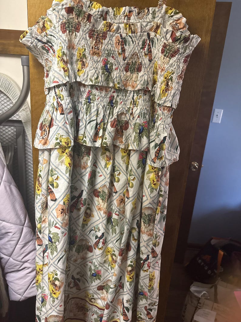 May include: A white dress with a floral pattern featuring birds, flowers, and fruit. The dress has a smocked bodice and a tiered skirt. The colors include yellow, orange, green, and blue. The dress has short sleeves and a midi length.