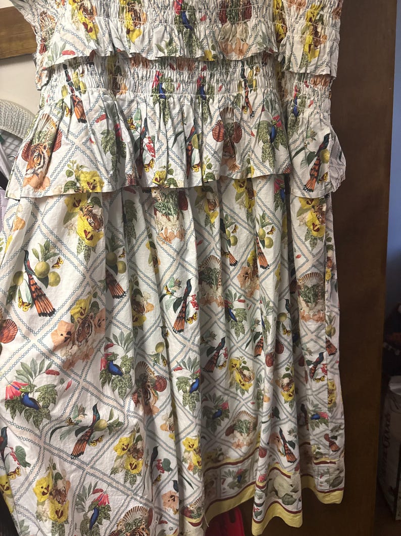May include: A white dress with a floral and bird print. The dress has a tiered design with ruffled details and elasticated sections. The pattern includes tigers, birds, and flowers in various colours, set against a diamond grid.