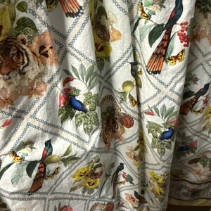 May include: White fabric featuring a repeating pattern of tigers, birds, butterflies, and flowers. The design includes a diamond grid with a chain-like border. The colour palette features shades of beige, green, yellow, and orange, creating a vibrant and detailed textile.