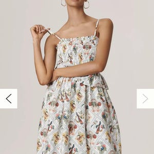 May include: A white sundress with a floral pattern featuring birds and flowers. The dress has a smocked bodice and spaghetti straps. The skirt is knee-length with a yellow hem.