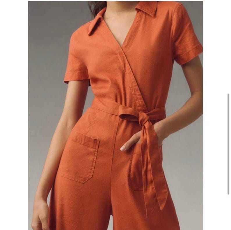 May include: An orange jumpsuit with short sleeves, a collared neckline, and a wrap-style front. The jumpsuit features a tie belt at the waist and two front pockets. The fabric appears to be a lightweight linen or cotton blend.
